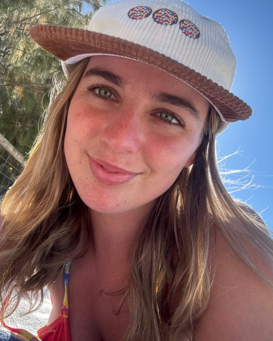 A woman smiling for a selfie on a sunny beach, wearing a white ribbed baseball cap with a brown brim and three colorful embroidered donut motifs on the front, along with a red bikini top.