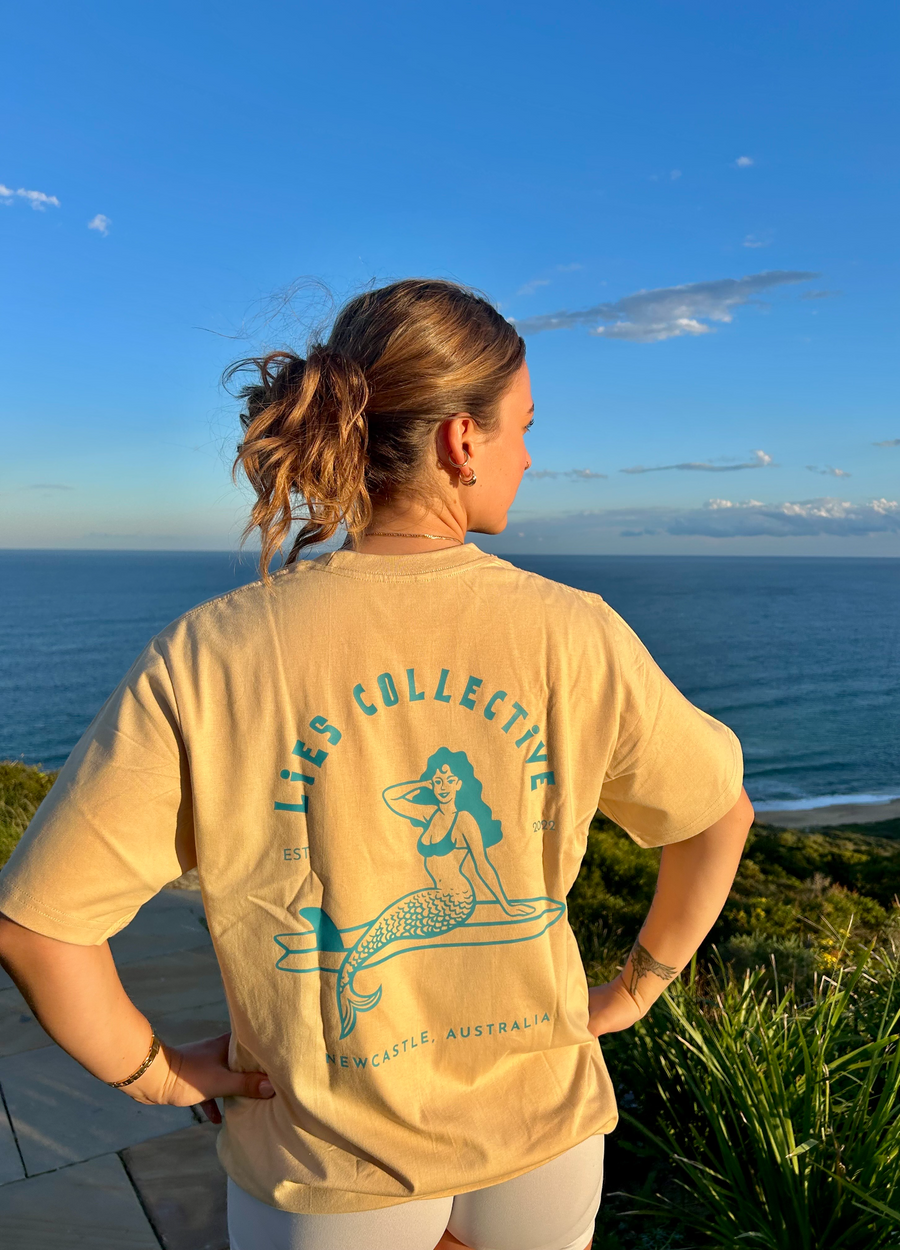 Beige short-sleeve tee with mermaid surfing graphic centered on back, crew neck